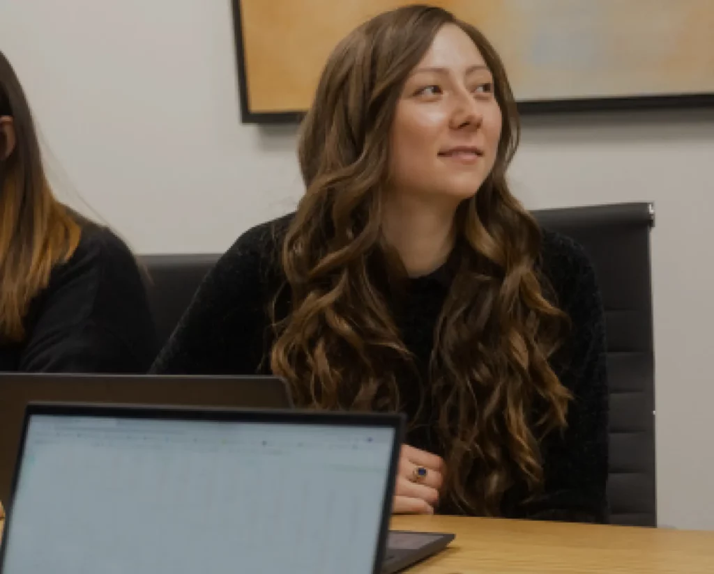 A woman sits at a desk. She has a laptop in front of her, but she is looking to the left.