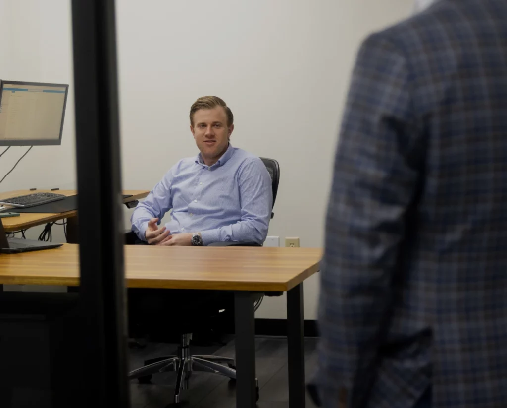 A CPA in a dress shirt sits behind a desk. He is speaking to someone directly in front of the camera.