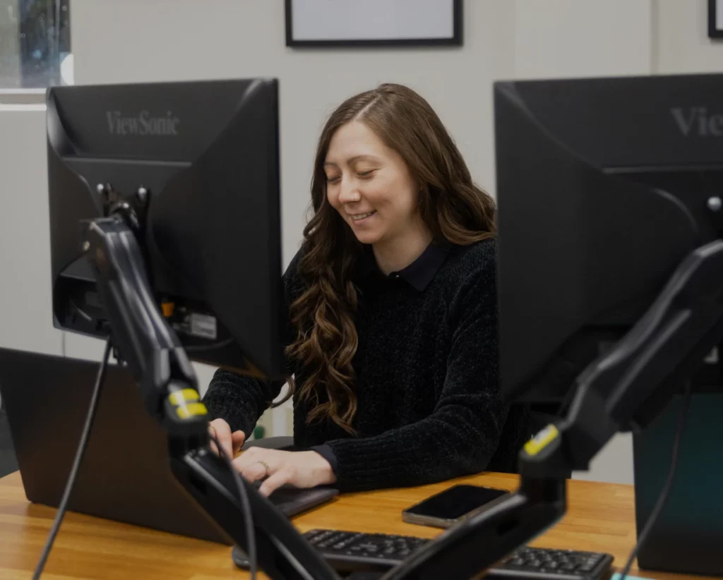 A CPA types on a laptop, smiling and looking down. There is an external monitor in front of her.